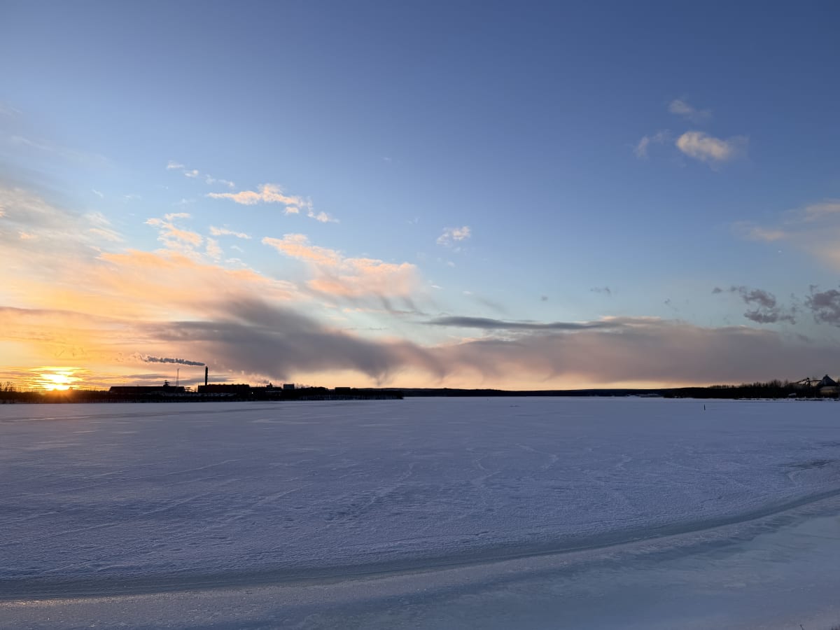 Bilden visar ett snötäckt sjö- eller havslandskap under en solnedgång eller soluppgång. Horisonten är låg, och den nedre delen av bilden domineras av is och snö med några mörkare områden som kan vara vatten eller tunn is. I bakgrunden, vid horisonten