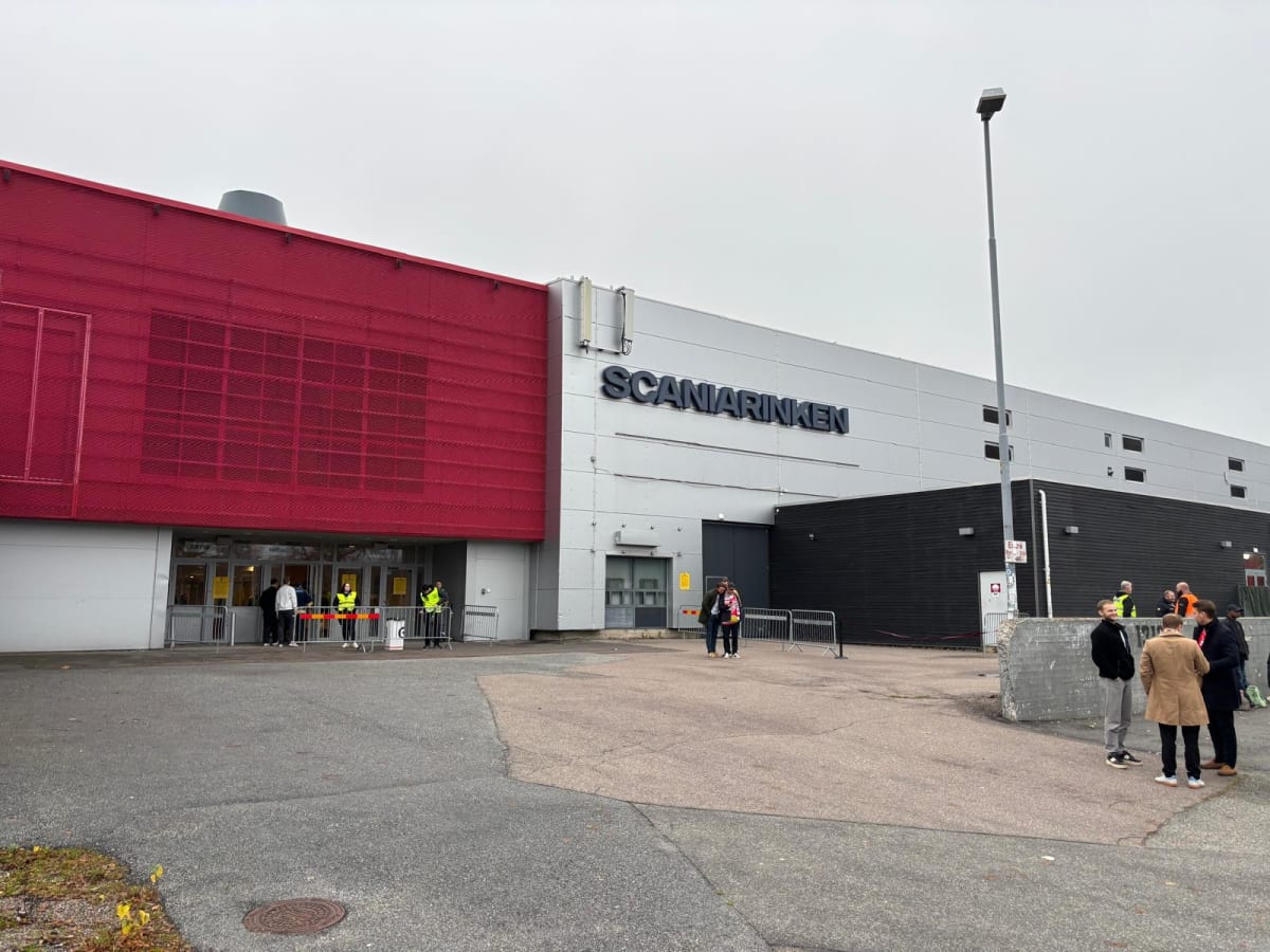 En utomhusbild föreställande en stor byggnad, uppdelad i sektioner med röd fasad till vänster och grå fasad till höger. På den grå fasaden syns texten "SCANIARINKEN" i stora svarta bokstäver. Framför byggnaden finns en asfalterad yta med flera person