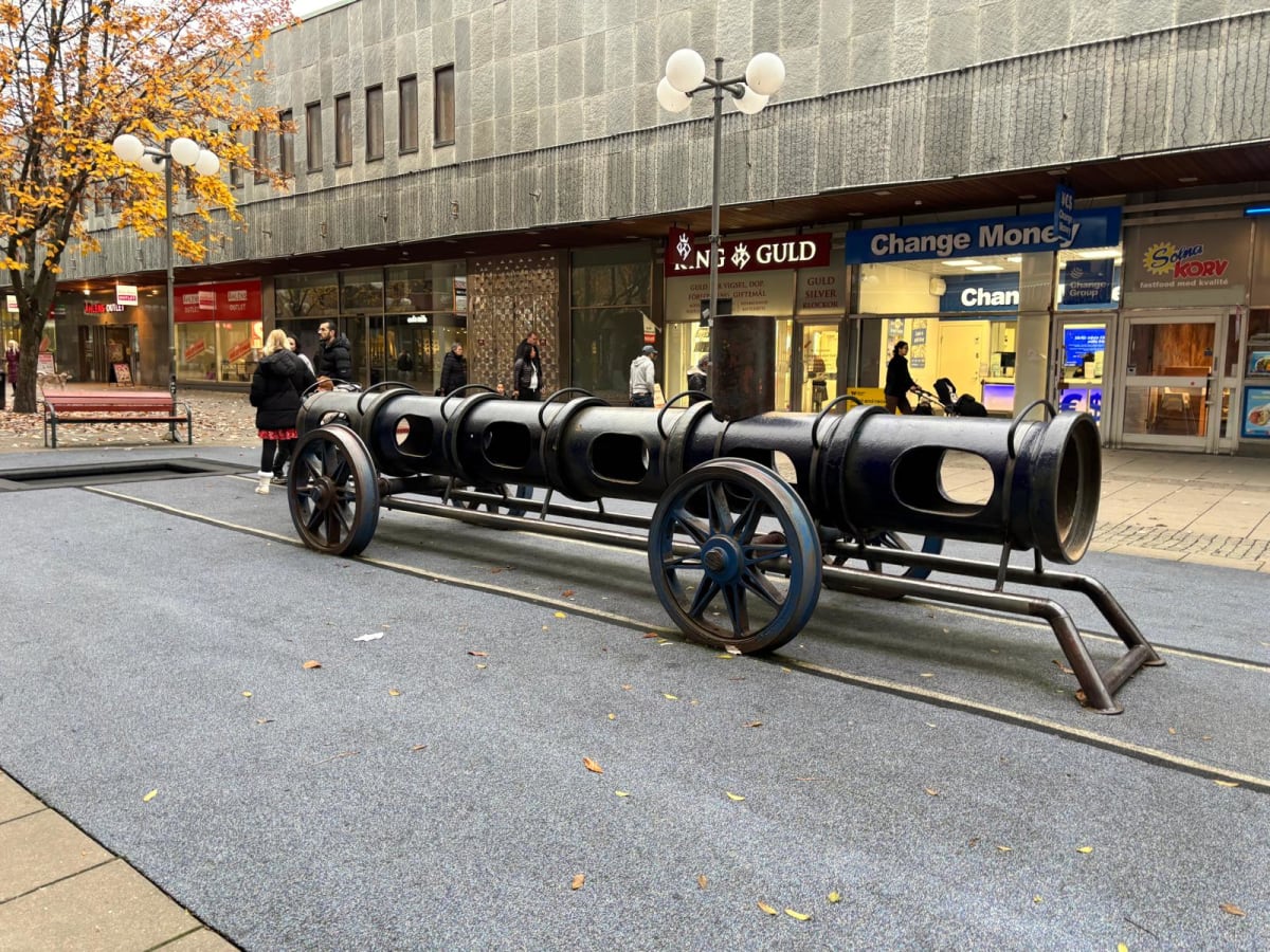 Bilden visar ett stadstorg eller en gågata med en stor, mörkfärgad leksakstågliknande skulptur i förgrunden, som är placerad på ett grått underlag. Skulpturen har flera cylindriska sektioner med öppningar och blåmålade hjul. I bakgrunden syns kommers