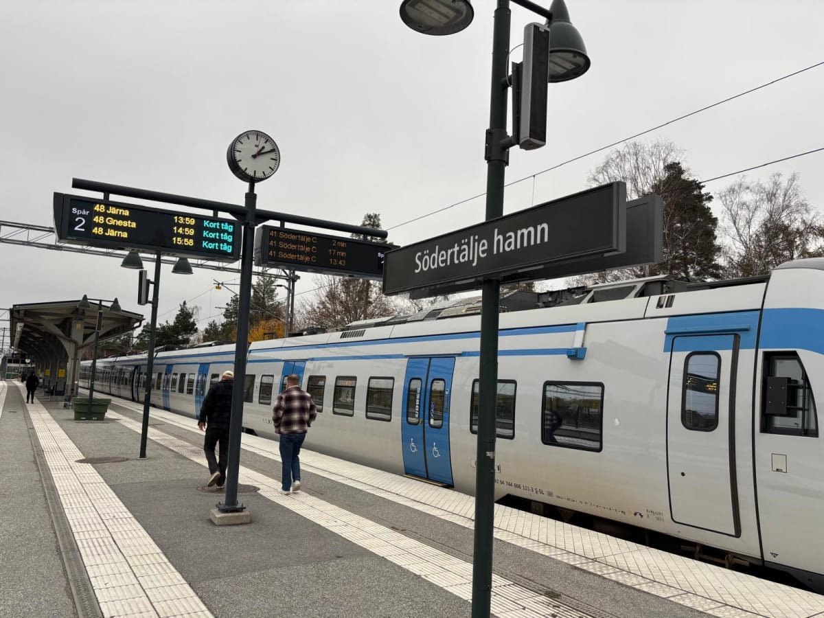 Bilden visar en järnvägsstation utomhus under dagtid med en tågvagn parkerad vid perrongen. Tåget är vitt med blåa detaljer och har fönster längs sidan. På perrongen syns flera informationsskyltar. En stor skylt indikerar "Södertälje hamn". En annan