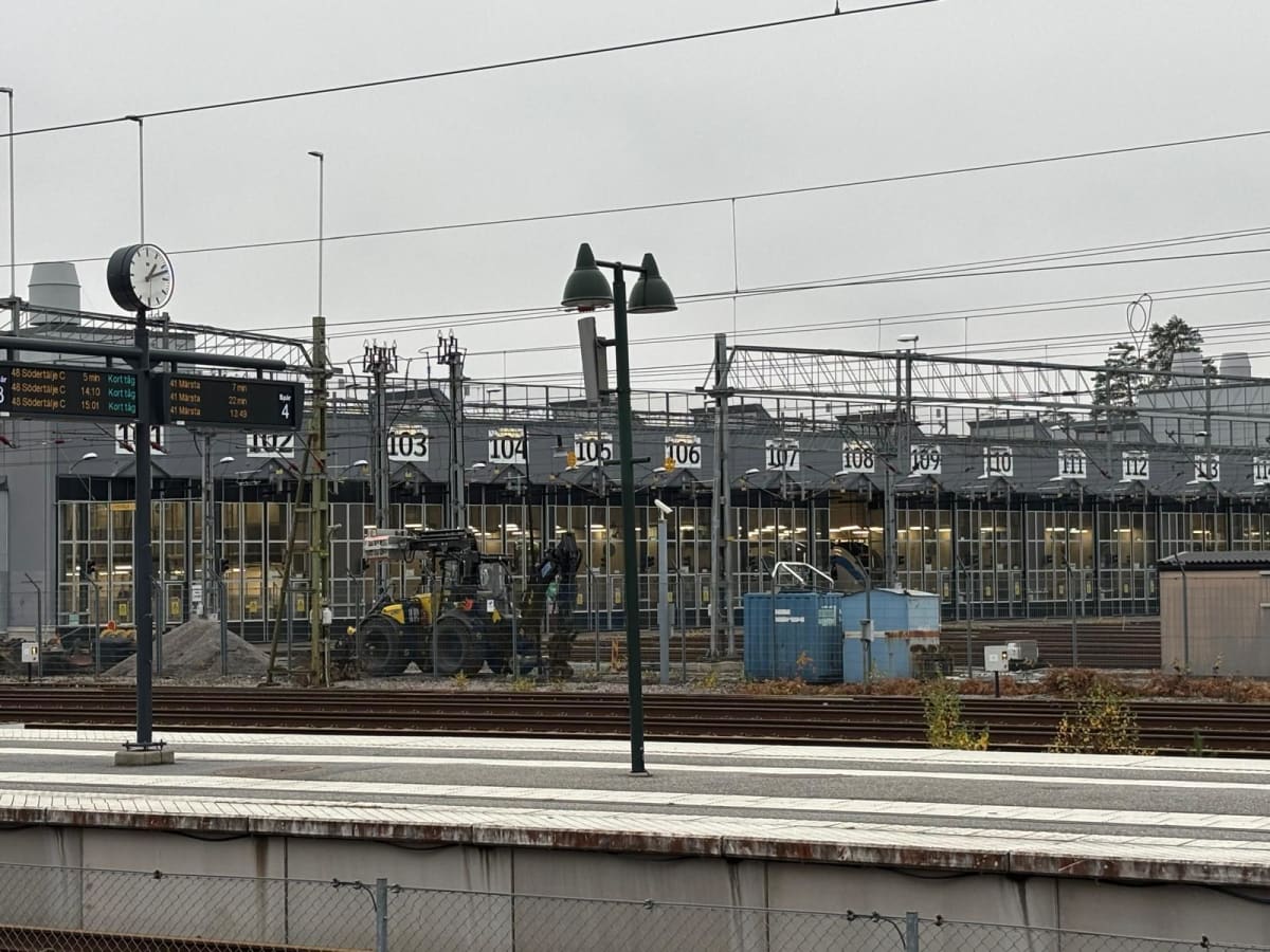 Bilden visar ett tågspår och en plattform på en järnvägsstation. I bakgrunden syns en stor byggnad med många garageportar, numrerade 103 till 114. Framför byggnaden finns ett flertal tågspår. Ett gult grävfordon med en lyftarm står vid ett av spåren.