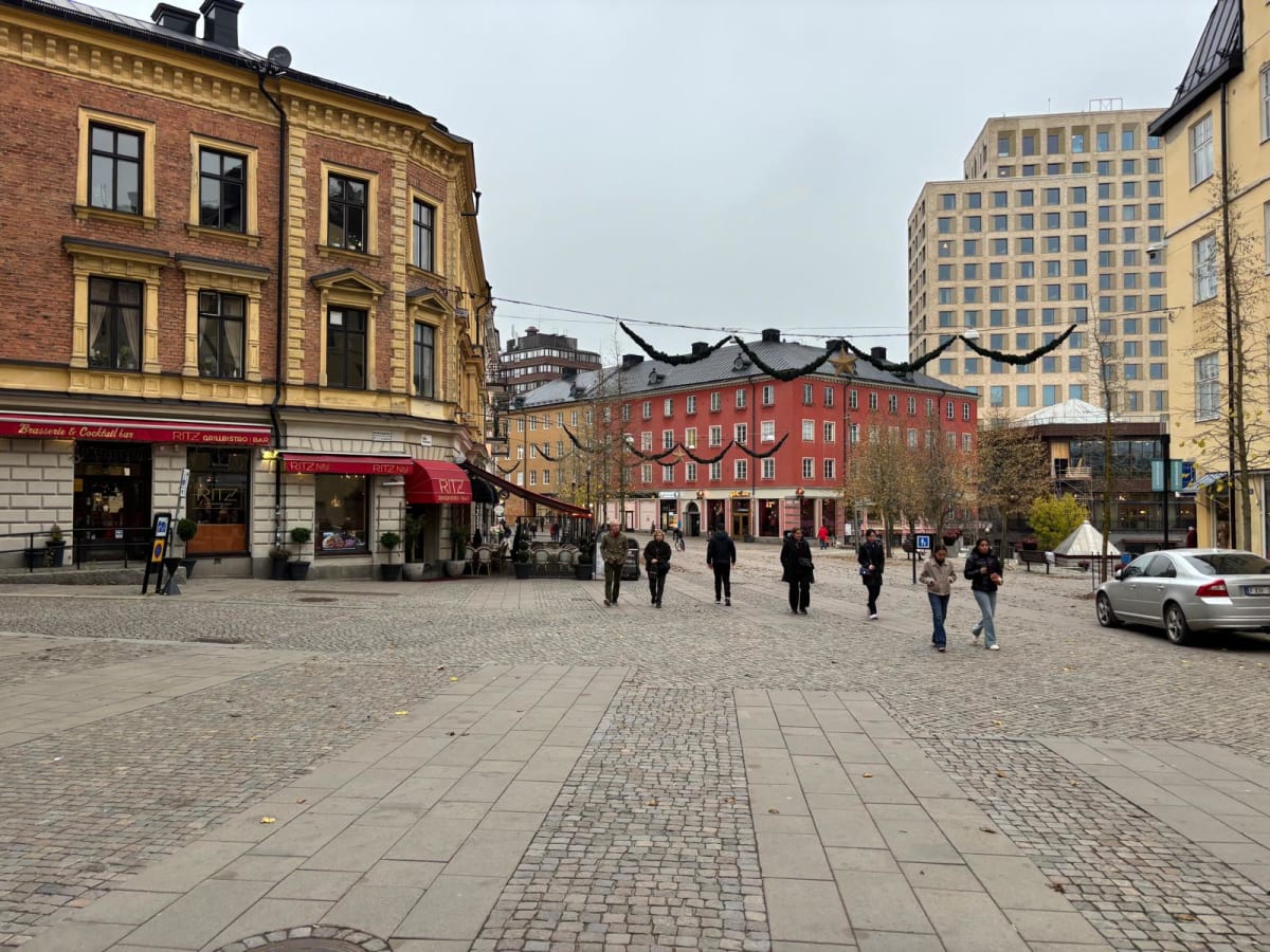 Bilden visar ett torg eller en öppen plats i en stad. I förgrunden syns gatsten och gångbanor. På vänster sida dominerar en tegelbyggnad med gula detaljer och svarta fönsterkarmar. Byggnaden inrymmer en "Brasserie & Cocktail bar" och "RITZ GRILLBISTR