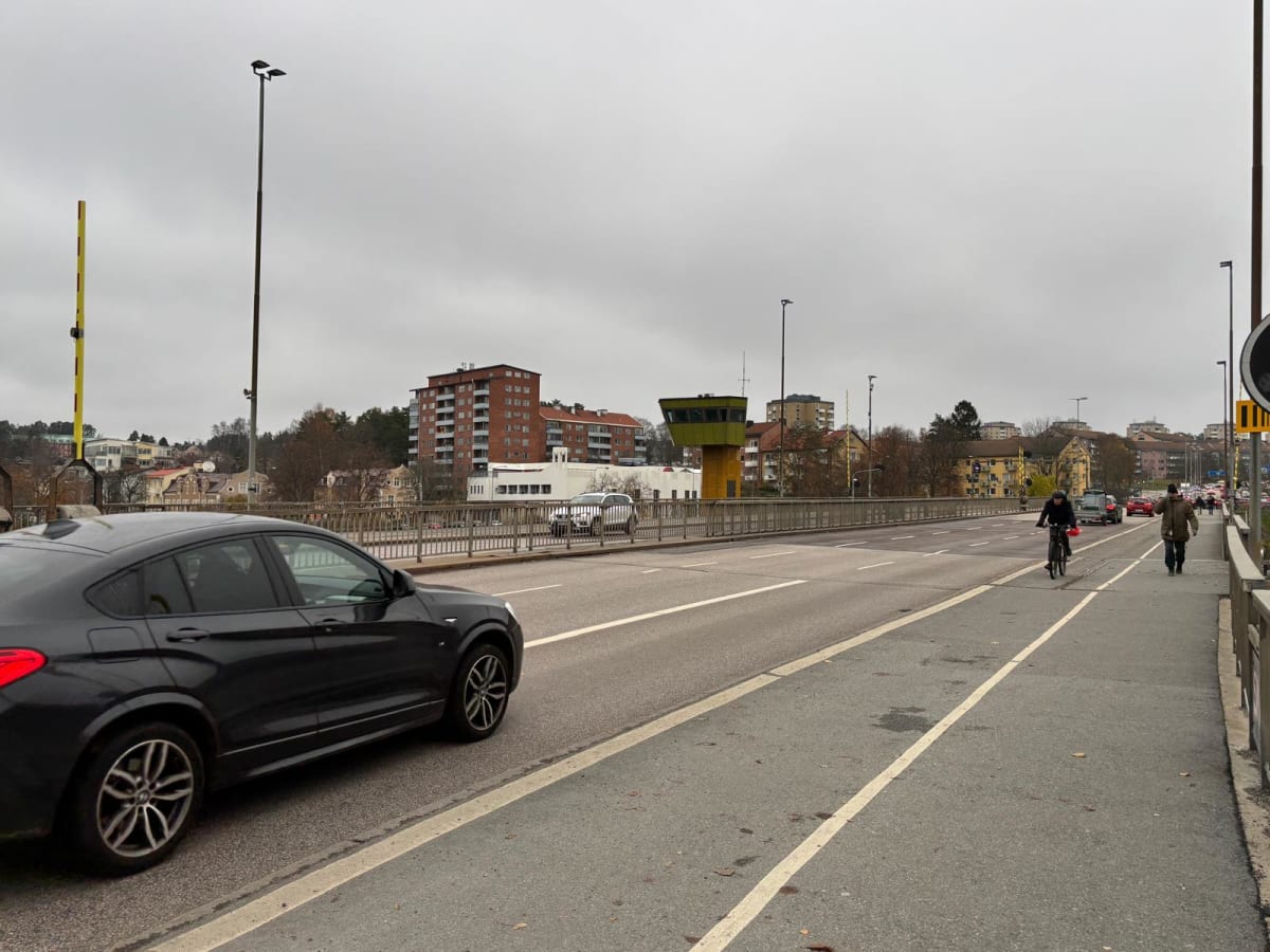 Bilden visar en vägbro med flera körfält, en cykelbana och en gångbana under en molnig himmel. En svart bil syns tydligt i förgrunden, på väg bort från kameran i det vänstra körfältet. Längre fram på bron syns en person som cyklar och en person som g