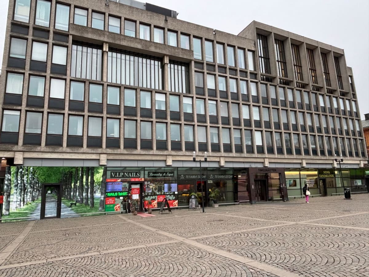 En bild tagen utomhus i ett stadsmiljö, föreställande en stor, rektangulär byggnad med en fasad av betong och många rektangulära fönster. Byggnaden dominerar bilden. På markplan finns flera butiker och verksamheter, inklusive 'V.I.P NAILS Beauty & Sp