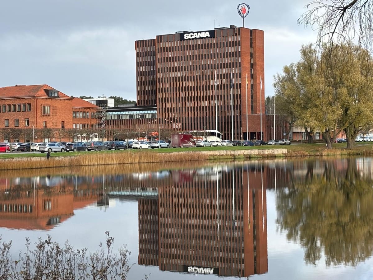 En stor, mörkbrun byggnad med texten "SCANIA" och en Scania-logotyp på taket står i centrum av bilden. Byggnaden reflekteras tydligt i en stilla vattenyta framför den. Till vänster om den stora byggnaden syns delar av en lägre, röd tegelbyggnad. En r