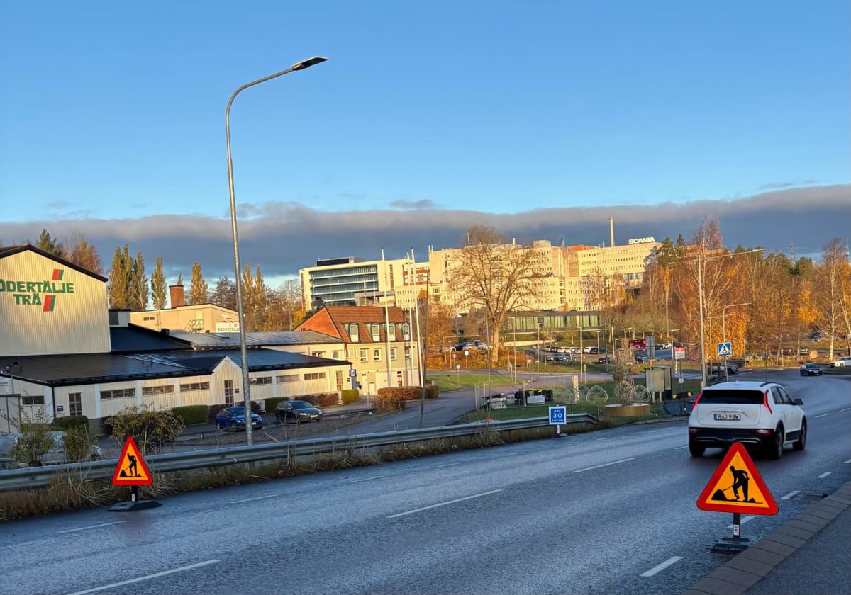 Bilden visar en vägsträcka från fågelperspektiv under dagtid. I förgrunden syns en vägbana som ser ut att vara våt, eventuellt från regn nyligen. På vägbanan finns två gul-röda varningsskyltar som indikerar vägarbete, med en figur som arbetar med en