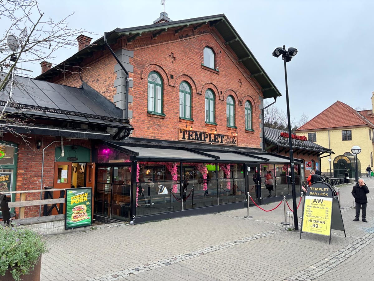En bild visar en tegelbyggnad med ett café och bar i förgrunden. Fasaden är av rött tegel med mörkgröna fönsterluckor och takfot. På bottenvåningen finns en inglasad uteservering dekorerad med rosa blommor, och ovanför den syns skylten 'TEMPLET Bar &