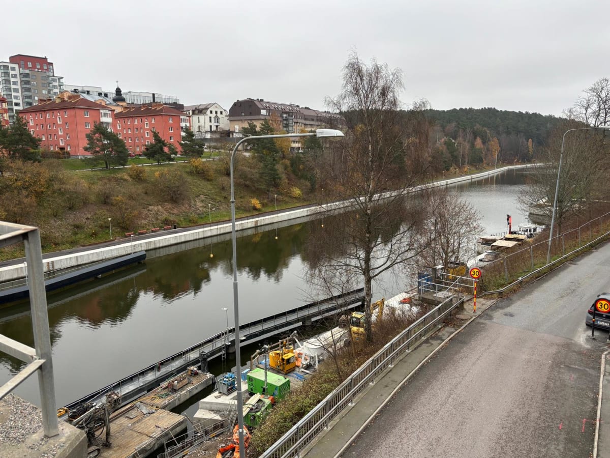Bilden visar ett landskap taget uppifrån med en kanal eller flod i fokus under en molnig himmel. Längs floden syns på ena sidan bostadsbyggnader i rött och en högre modern byggnad i ljusa färger, omgivna av vegetation som visar tecken på höst. På den