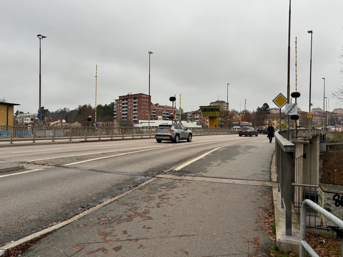 Bilden visar en bro med trafik en molnig dag. På vägen syns flera bilar, inklusive en ljusgrå SUV i förgrunden som kör bort från kameran, och en mörkare bil och en buss längre bort. En person går på gångbanan till höger i bild. Längs brons sidor finn