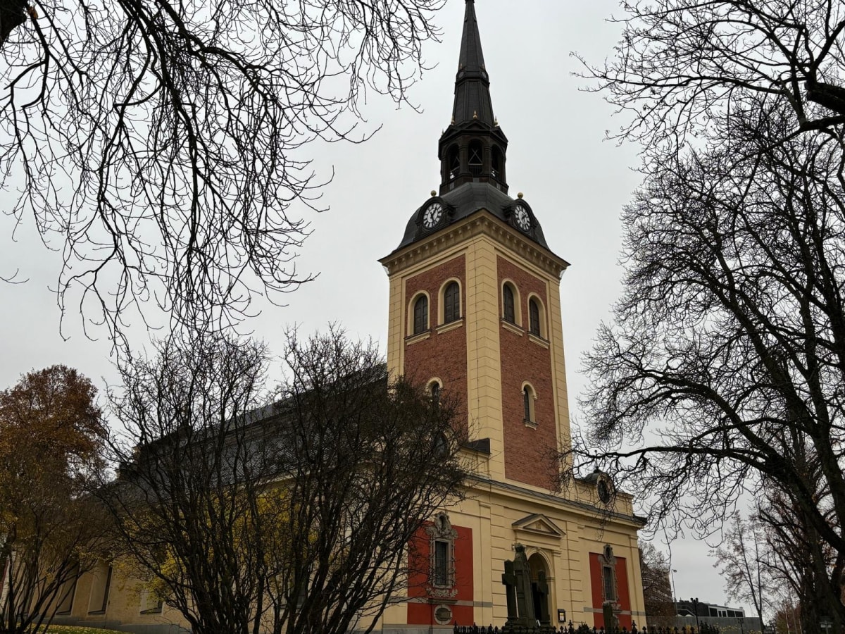 Bilden visar fasaden och tornet av en kyrka, sedd delvis skymd av nakna trädgrenar i förgrunden. Kyrktornet har en spetsig spira med en kupolformad bas och klockor på varje sida. Tornet är uppbyggt med tegel och gula fasaddetaljer. Huvudbyggnaden är