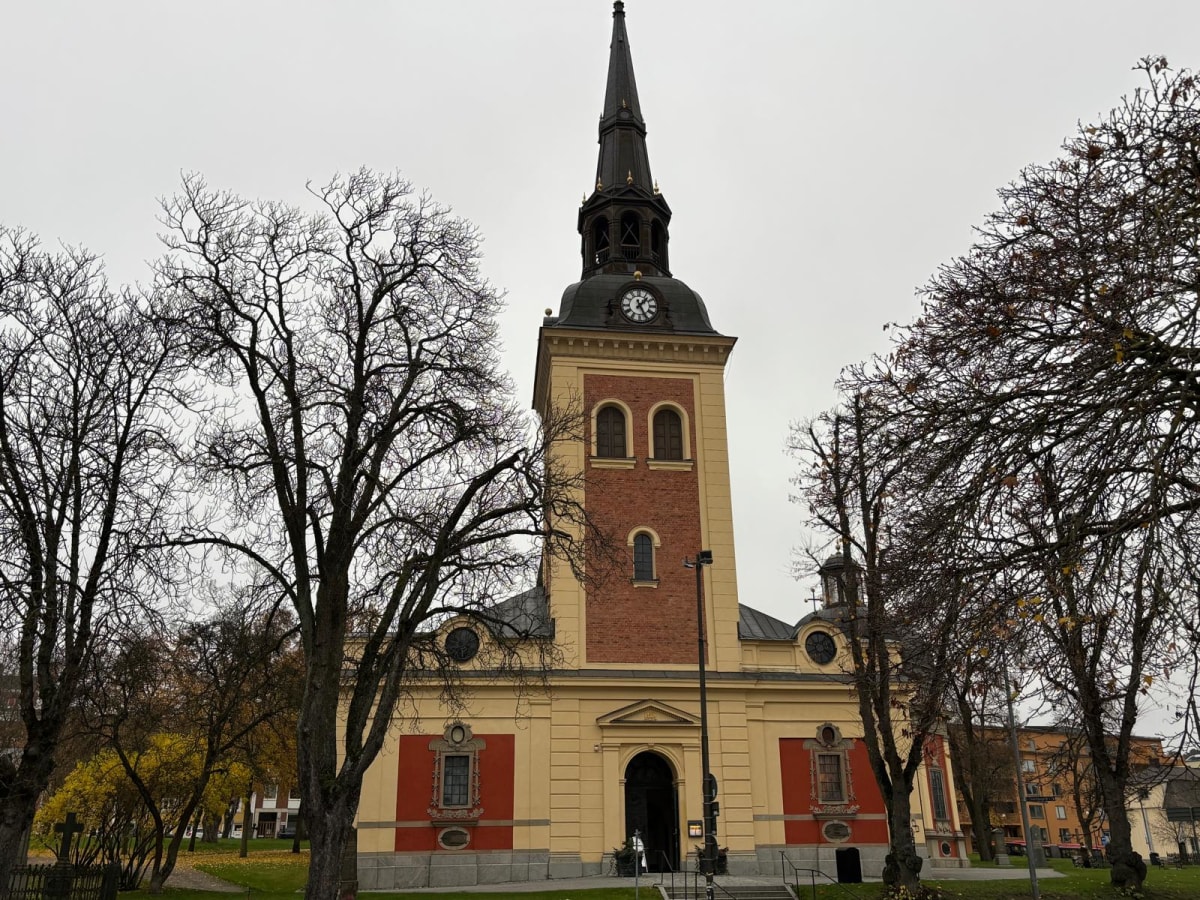 Bilden visar fasaden på en gul och röd kyrkobyggnad med en hög spira, tagen under dagtid. Kyrkan har en stor brun spira med ett klocktorn som visar visare och siffror, samt ett flertal fönster i rundad bågform. Delar av kyrkan är täckta av tegel, med