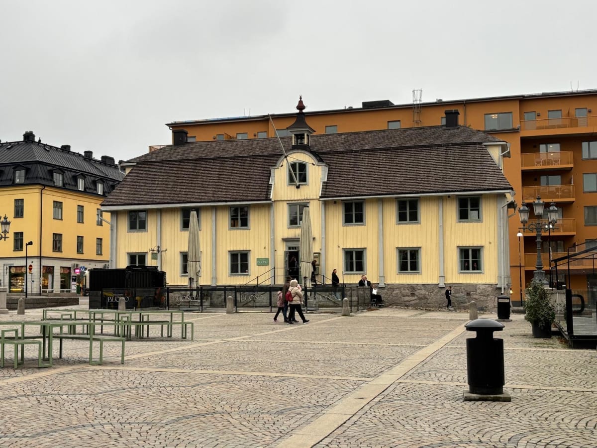 Bilden visar ett torg eller en öppen plats framför en stor, gul byggnad med brunt tak. Byggnaden har många fönster med mörkare ramar och en liten takkupa med flaggstång i mitten. Två stora hopfällda parasoller syns på fasaden. I förgrunden till vänst