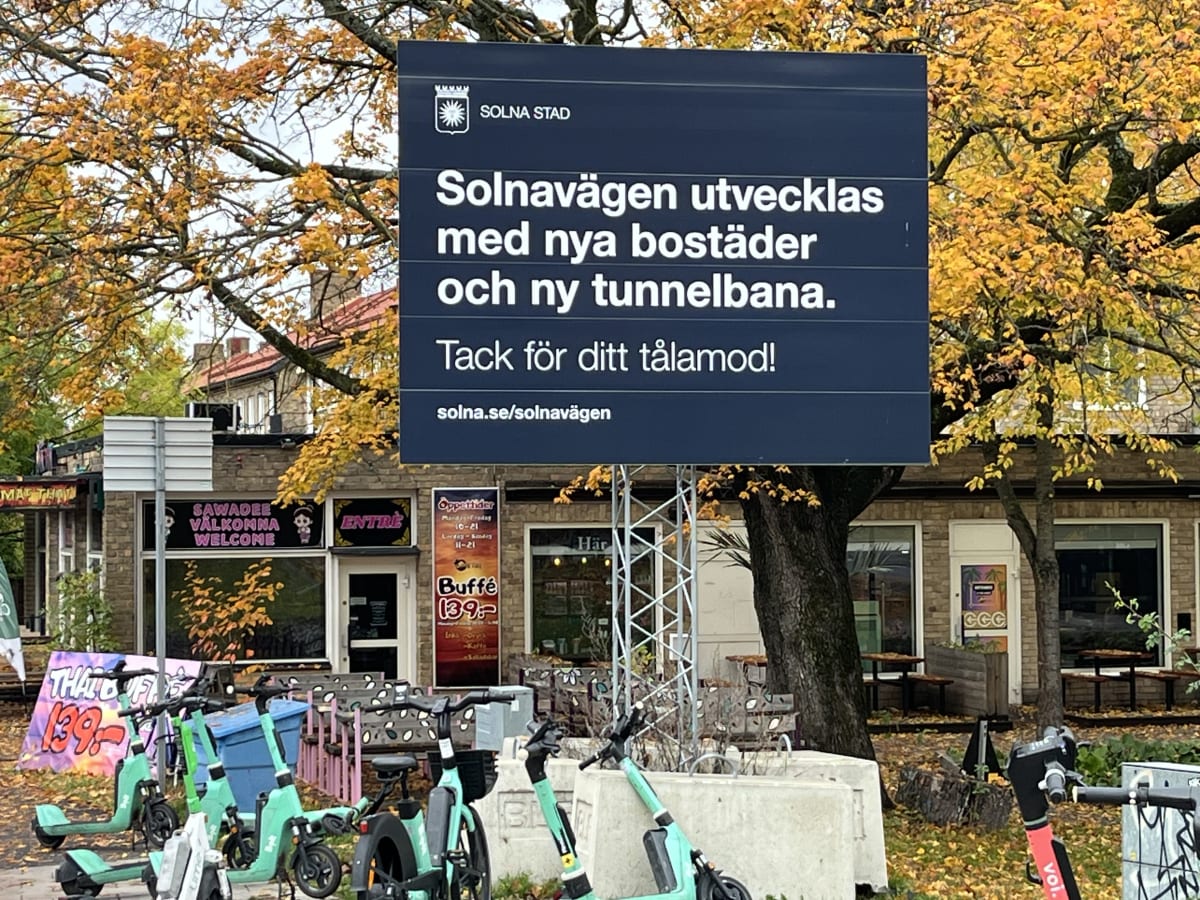 Bilden visar en gatuscen under hösten med löv på träden i gula och orangea nyanser. I förgrunden står ett flertal elsparkcyklar och en elcykel parkerade. Centralt i bilden dominerar en stor blå skylt från Solna Stad med texten "Solnavägen utvecklas med nya bostäder och ny tunnelbana. Tack för ditt tålamod!" samt webbadressen "solna.se/solnavägen". Bakom skylten syns delar av en tegelbyggnad med fönster och dörrar samt reklam för en restaurang, inklusive en banderoll med texten "SAWADEE VÄLKOMNA WELCOME" och en skylt som annonserar "Buffé 139:-". Hela scenen är utomhus och det är dagsljus.