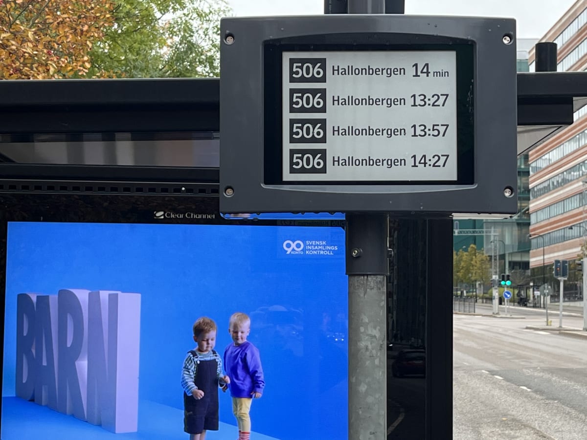 Bilden visar en digital informationstavla för busstider och en reklamskärm på en busshållplats. Överst på bildskärmen visas avgångstider för busslinje 506 till Hallonbergen, med den nästa bussen om 14 minuter och därefter fasta tider klockan 13:27, 1