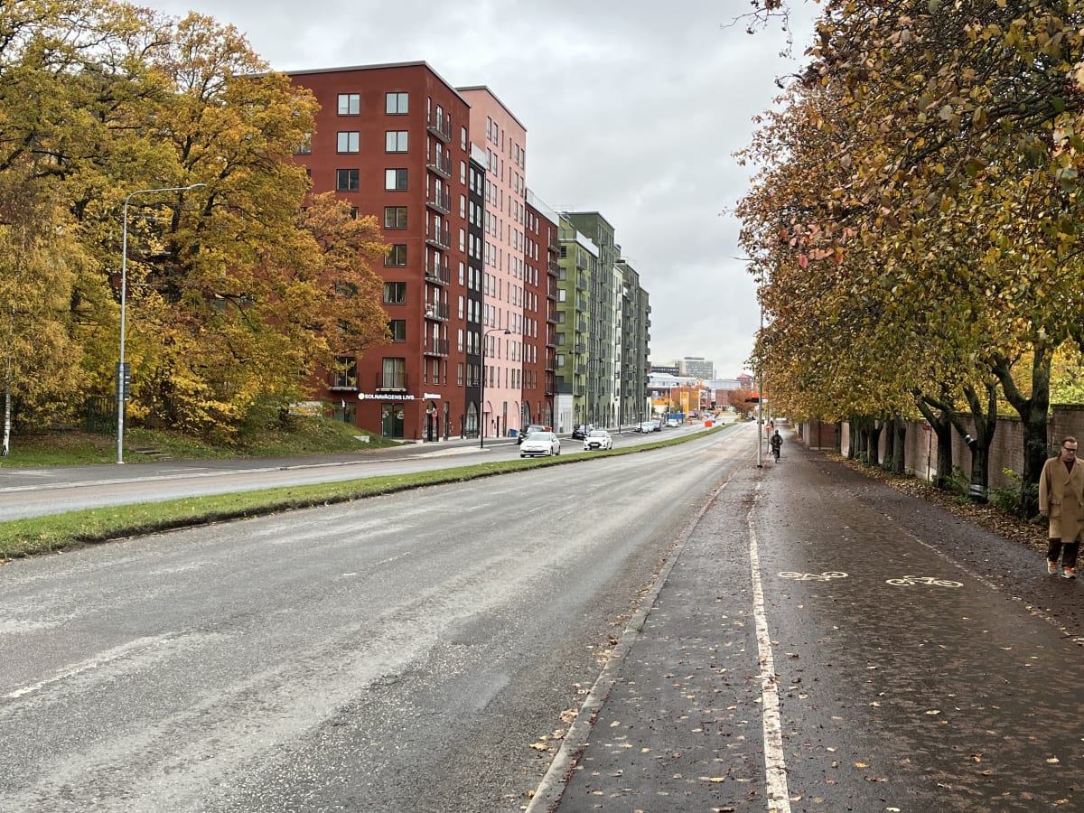 Bilden visar en gata i ett stads- eller förortsområde under dagtid med höstlövsfärgade träd. Till vänster om bildens mitt finns en vägbana för motorfordon, åtskild av en gräsremsa från en gång- och cykelbana till höger. Vägen är våt, vilket antyder r