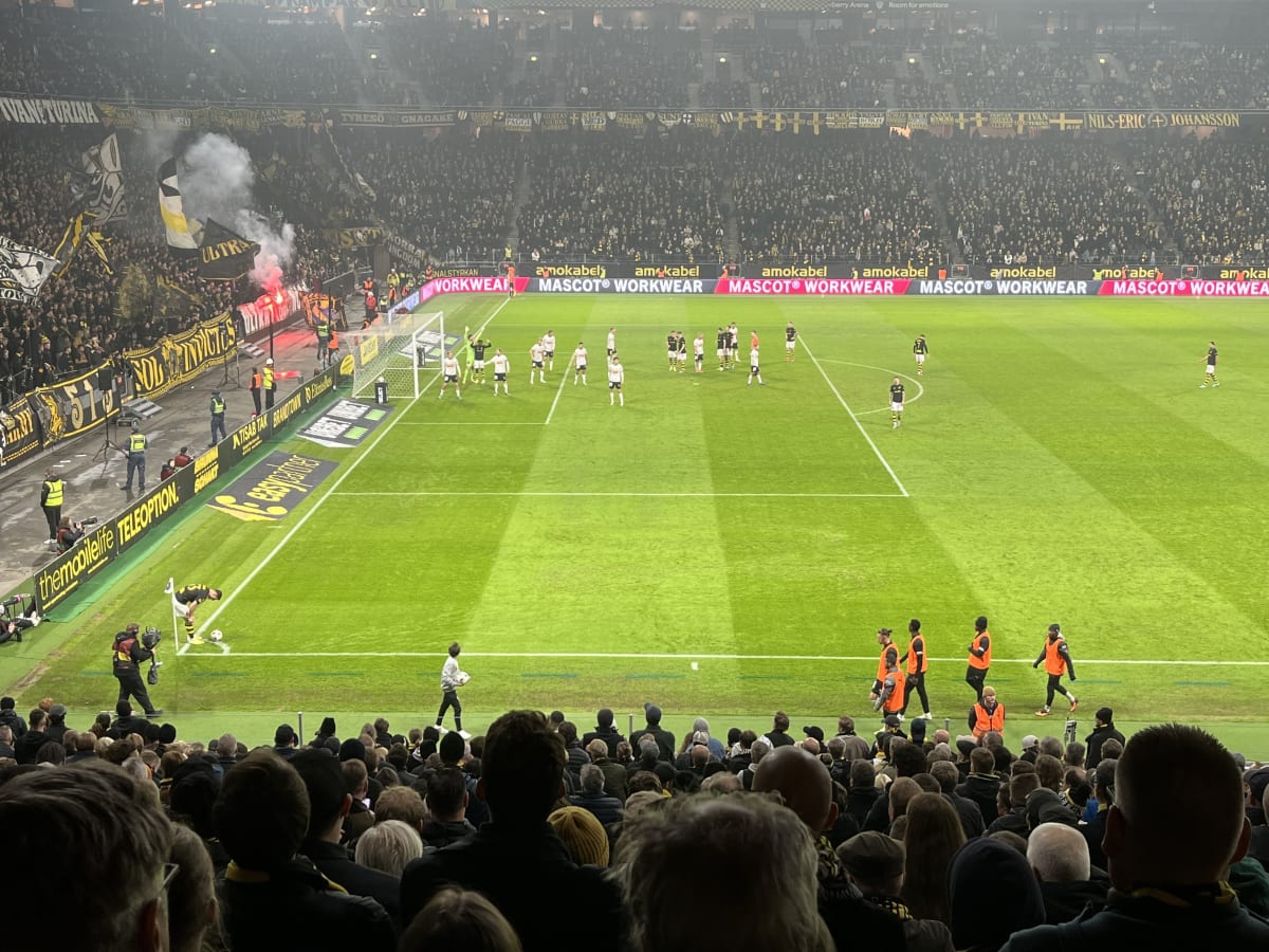 En vidvinkelbild tagen från en läktare på en fotbollsarena. På planen spelas en fotbollsmatch, med spelare i vita respektive svarta/gulrandiga dräkter. En spelare i svart/gulrandig dräkt förbereder sig för en hörnspark. Vid ena kortsidan av planen sy