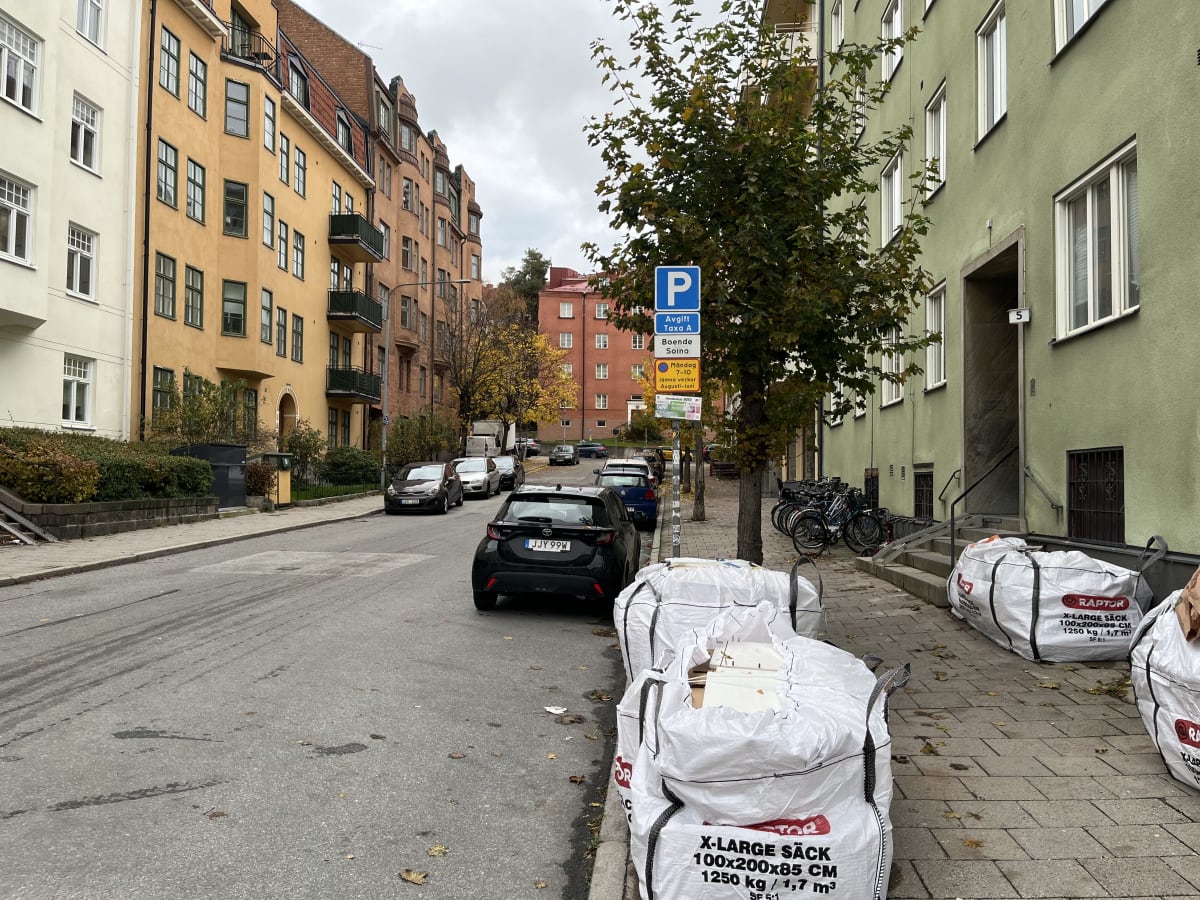 Bilden visar en gata med parkering och flerbostadshus på sidorna under en molnig dag. På vänster sida av gatan syns en rad parkerade bilar, varav en svart bil med registreringsnummer JJY 99W står närmast kameran. Till vänster om gatan står en gul byg
