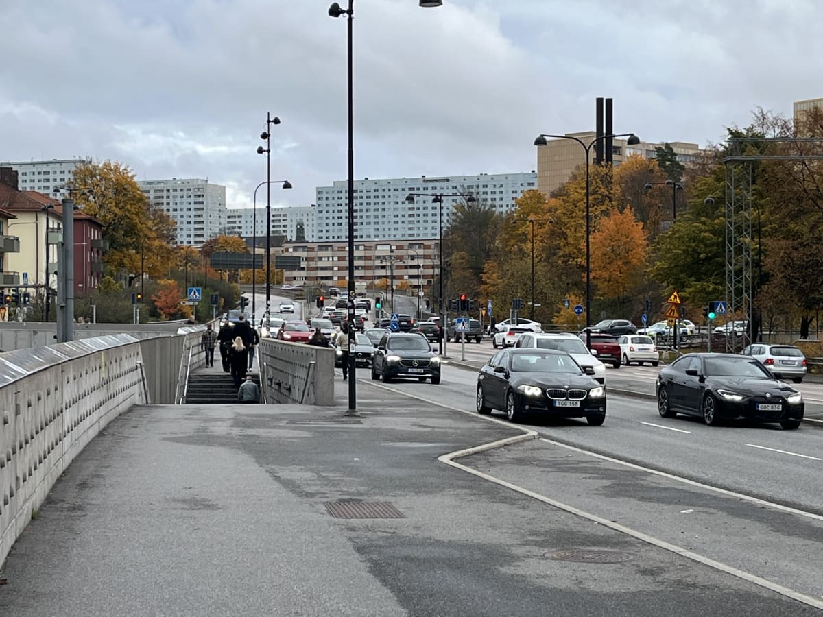 Bilden visar en stadsmiljö med en bred väg och flera bilar i rörelse, samt fotgängare på en trottoar och en gångbro. Till vänster i förgrunden finns en gångbro eller övergång med trappa, där flera personer går. Vägen är fylld med bilar, de flesta mör