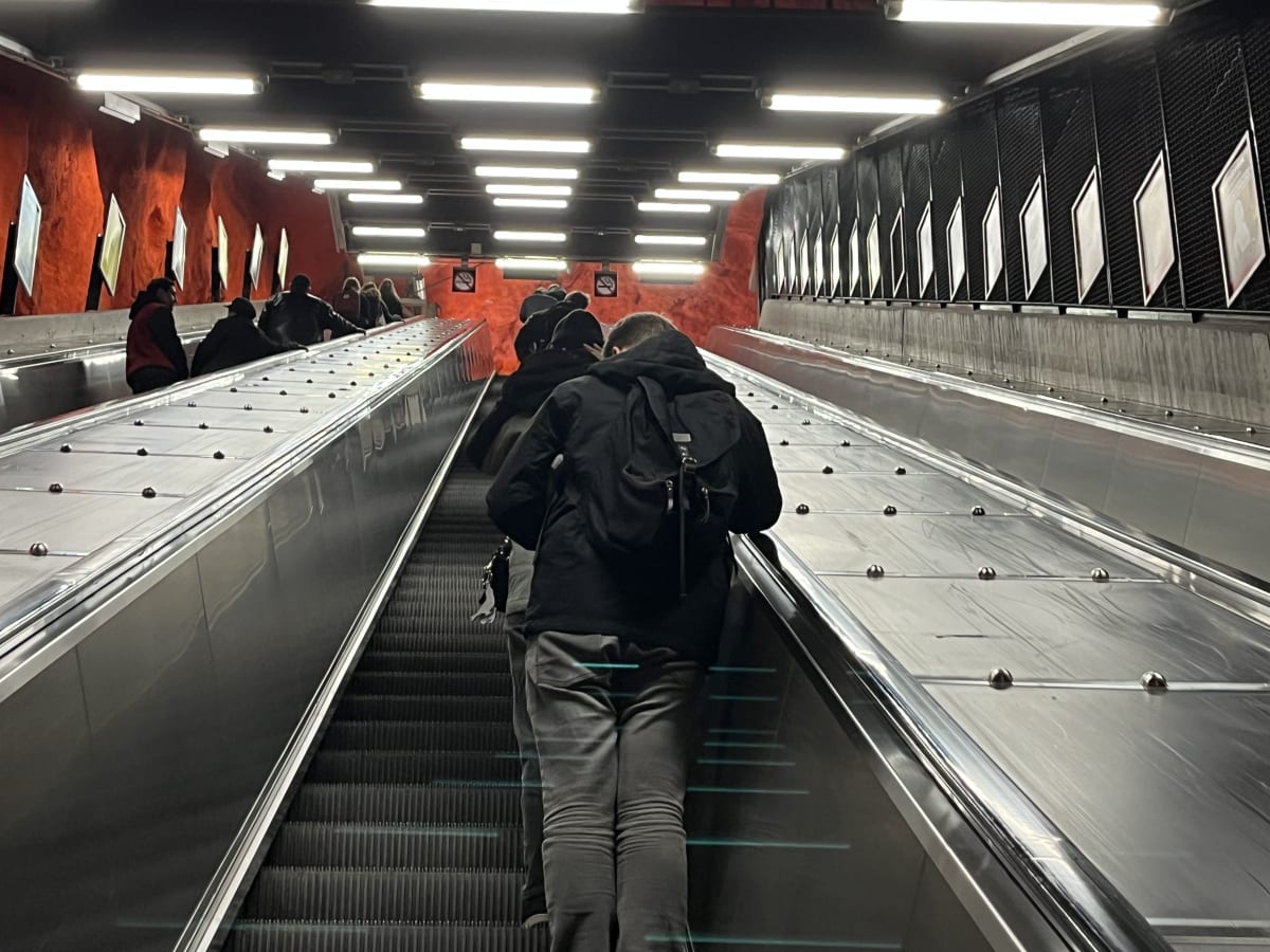 Bilden visar människor som färdas uppför två rulltrappor i en tunnelbanestation eller liknande offentlig transportmiljö. Rulltrapporna är av metall och har ett modernt utseende. Väggarna bredvid rulltrapporna har en distinkt orange, ojämn textur på ena sidan, som liknar klippa eller grottvägg, och är upplysta av ett flertal rektangulära lysrörslampor i taket. På den orangeröda väggen sitter det också inramade, ljusa paneler. Den andra sidan av rulltrappan har en mörkare vägg med svarta galler och liknande ljusa paneler. Flera personer, huvudsakligen vuxna, syns bakifrån i rulltrapporna, de flesta bär mörka kläder, inklusive en person i förgrunden med en ryggsäck. Några informations- eller varningsskyltar syns också på den orangeröda väggen.