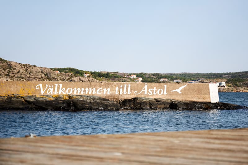 Bilden visar ett marint landskap under en klarblå himmel. I förgrunden syns en del av en träbrygga. Mellan bryggan och horisonten sträcker sig ett vattenområde med små vågor. I mitten av bilden, placerad i vattnet, finns en betongkaj eller ett fundam