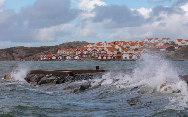 Bilden visar ett landskap med kraftiga vågor som slår mot en stenpir i förgrunden. Vattnet verkar vara upprört och skummande. I bakgrunden, på andra sidan vattnet, syns ett samhälle med många hus. De flesta husen har röda tak och några är vita eller