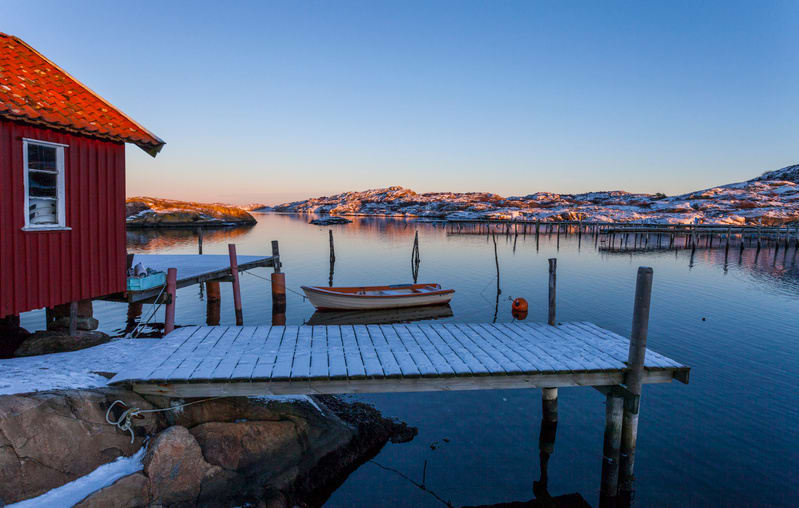 En bild visar en vinterlandskap med ett rött sjöbod till vänster, dess tak är tegelrött och det finns ett vitt fönster. Intill sjöboden ligger en träbrygga nedsläppt i vattnet, med ett tunt lager snö som täcker dess yta. I vattnet flyter en liten, lj