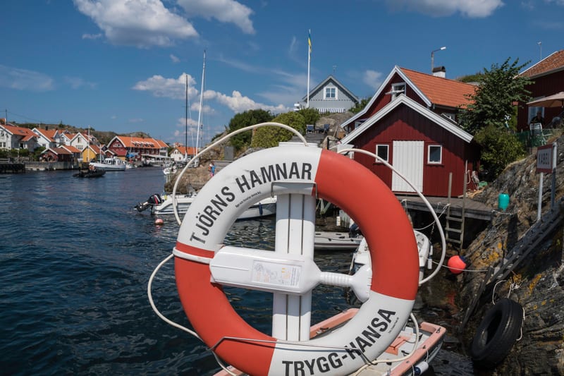 Bilden visar ett landskap med en hamn och hus. I förgrunden syns en röd och vit livboj med texten "TJÖRNS HAMNAR" och "TRYGG-HANSA". Bakom livbojen är vatten med båtar, inklusive en liten jolle och andra större båtar. I bakgrunden finns flera hus, me