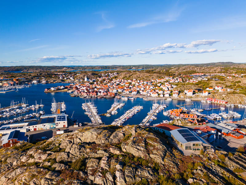 Bilden visar ett flygfoto över en tätbebyggd kuststad eller ett fiskeläge med många byggnader med röda tak. Staden är omgiven av ett kuperat, klippigt landskap med delvis sparsam vegetation. Centralt i bilden finns en stor hamn med ett stort antal bå