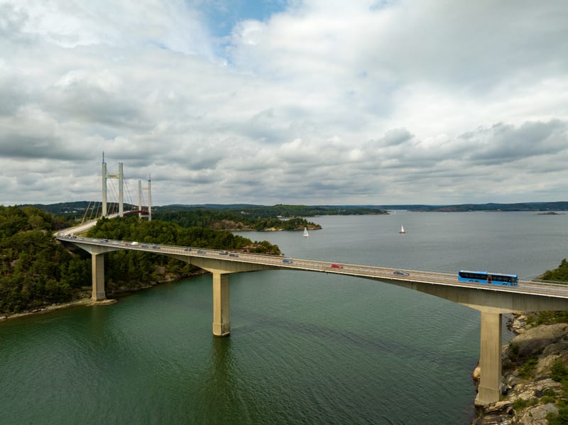 En vidvinkelbild tagen från ovan visar en bro som sträcker sig över vatten. På bron syns flera fordon, inklusive en blå buss i förgrunden och ett antal personbilar. Bron är kopplad till landområden på båda sidor, vilka är täckta med grön vegetation o