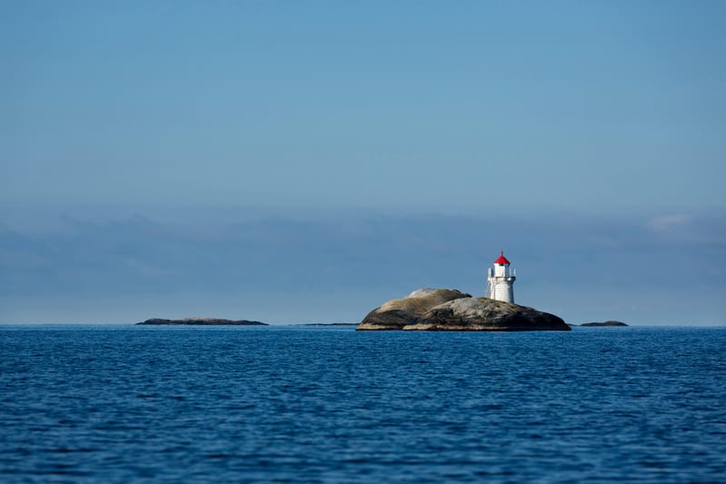 En bild som visar en vit fyr med ett rött tak placerad på en liten, klippig ö mitt i ett vattenområde. Fyren är mitt i bilden mot en blå himmel. Till vänster om den klippiga ön med fyren syns ytterligare en mindre klippformation som sticker upp ur va