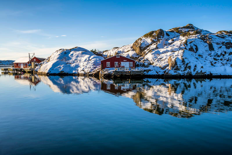 Bilden visar ett vinterlandskap med snötäckta klippor och byggnader vid ett lugnt vatten. Tre röda byggnader med vita detaljer, troligen sjöbodar eller hus, står på pålar eller direkt på klipporna vid vattnet. En av byggnaderna i förgrunden reflekter