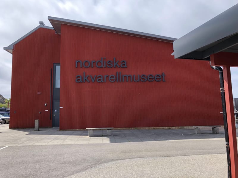 Bilden visar fasaden på en röd byggnad gjord av träpanel. På fasaden står texten "nordiska akvarellmuseet" i stora, mörka bokstäver. Byggnaden har ett grått tak med synliga takrännor. En dörr med glasparti är synlig i byggnadens vänstra del. Framför