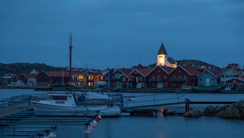 Bilden visar ett kustlandskap vid skymning eller gryning, med en by eller tätort belägen vid vattnet. I förgrunden syns en hamn med flera båtar förtöjda vid bryggor. Vissa båtar är vita och en har ordet "PIRRIANA" skrivet på sidan. Bakom hamnen sträc