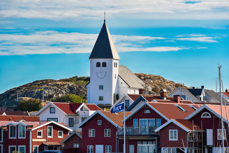 Bilden visar ett kustlandskap med en vit kyrka med ett mörkt pyramidformat tak och en leende ansiktsmålning på dess torn. Kyrkan är placerad på en kulle i bakgrunden, omgiven av bergliknande landskap med sparsam vegetation. I förgrunden syns flera rö