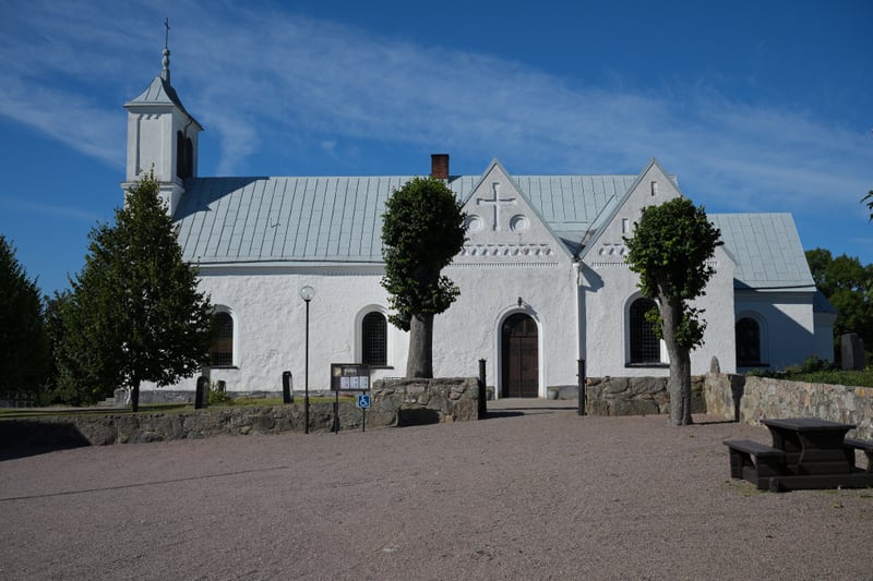 Bilden visar en vit kyrkobyggnad med ett ljust tak under en klarblå himmel med tunna vita moln. Kyrkan har en klockstapel med ett kors på västra sidan och flera gavlar mot syd. Fasaden är putsad i vitt och har välvda fönster med galler samt en mörkbr