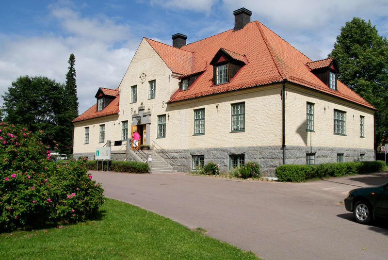 En stor, ljusgul byggnad med orange tegeltak och svarta skorstenar står i centrum av bilden under en molnig himmel. Byggnaden har flera fönster med vita karmar och mörkgröna spröjs. Fasaden är murad, med en grå stensockel. Vid huvudentrén, som har en