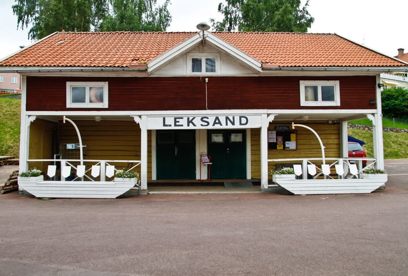 Bilden visar fasaden på en byggnad med en takkonstruktion i tegel och en front i träfinish. Byggnaden har en veranda med vita pelare och ett räcke som är utformat som akterskeppet på en båt med blomma. Mellan pelarna syns en gul träpanel med två dörr