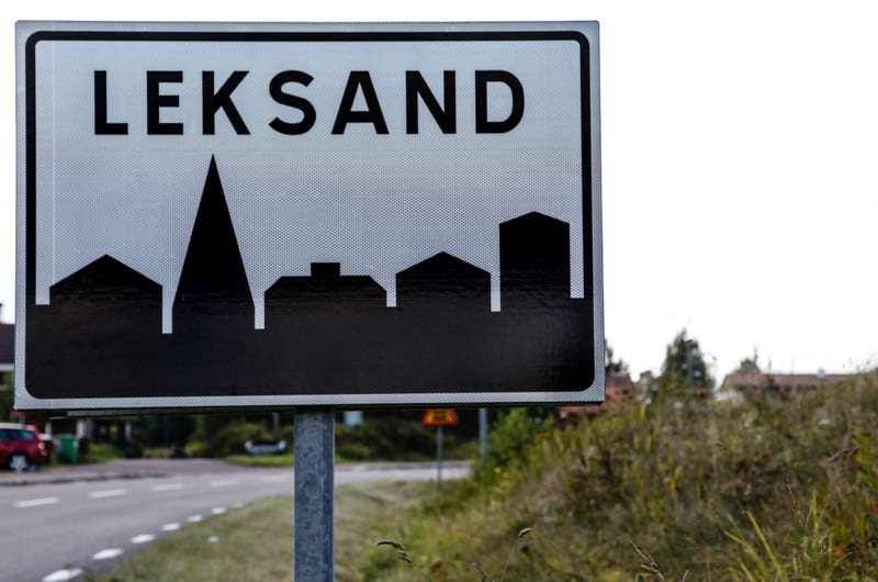 En vägskylt med texten "LEKSAND" i svarta versaler mot en gråvit bakgrund står i förgrunden. Under texten finns en svart silhuett av byggnader och en kyrkspira. Skylten är monterad på en metallstolpe. I bakgrunden syns en väg med vit körfältsmarkerin