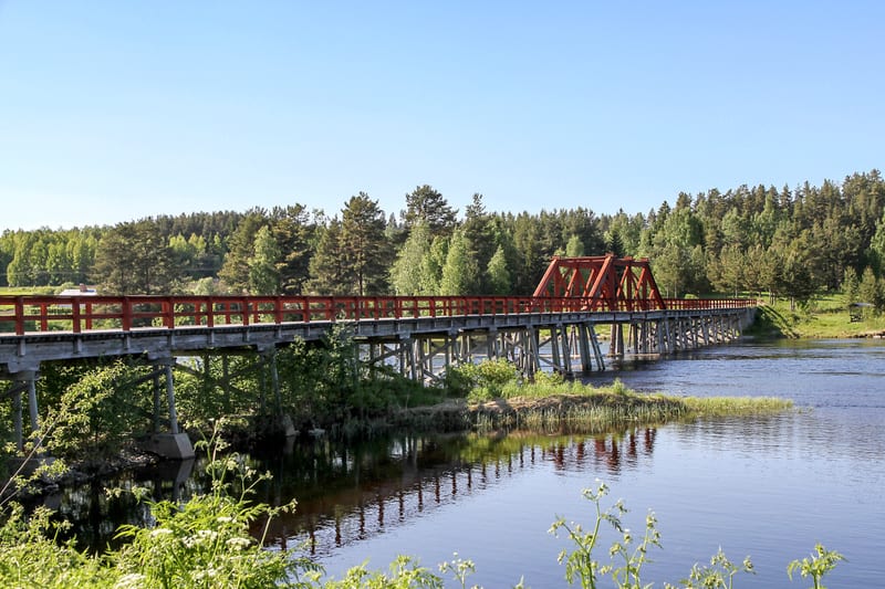 Bilden visar en lång bro i trä och stål som sträcker sig över en flod under klarblå himmel. Brobanan är träfärgad med ett rött räcke. En del av bron har en röd fackverkskonstruktion i stål. På vardera sidan av floden syns grönskande skog och vegetati