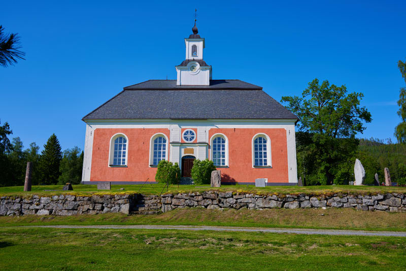 En rödskurad kyrkobyggnad med vittramade fönster och en svart takpapp mot en klarblå himmel. Kyrkan har en klockstapel med svart spira och vita urtavlor. Framför kyrkan finns en gräsklädd sluttning som övergår i en stenmur. Nedanför stenmuren syns en