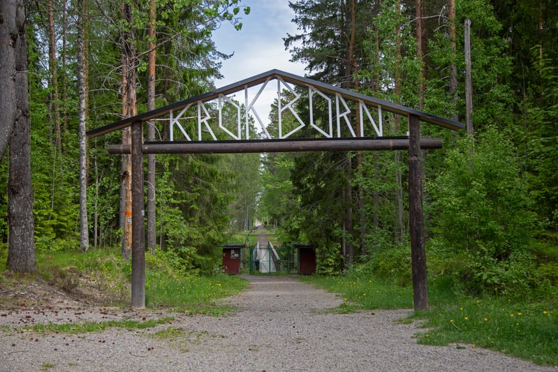 En bild tagen utomhus visar en grusväg som leder fram till en entréport med en skylt där det står "KROKBORN" i vita bokstäver mot en mörk bakgrund. Entréporten består av två vertikala trästockar som bär upp en horisontell trästock, över vilken det fi