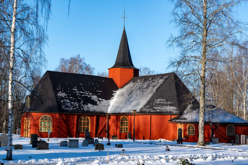 En röd träkyrka med svart tak syns i mitten av bilden, omgiven av ett snötäckt landskap med träd. Kyrkan har en spetsig takspira med en förgylld utsmyckning. Flera runda fönster med gula fönsterkarmar är synliga på kyrkans sidor. Taket är delvis täck