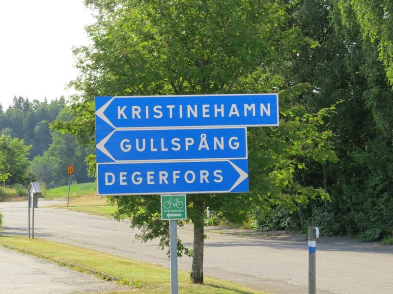 Bilden visar flera vägskyltar i ett soligt utomhusmiljö. Överst i bilden finns en blå vägskylt med vit text som pekar vänster och säger 'KRISTINEHAMN'. Under den finns en likadan skylt som pekar vänster och texten 'GULLSPÅNG'. Längst ner bland de blå