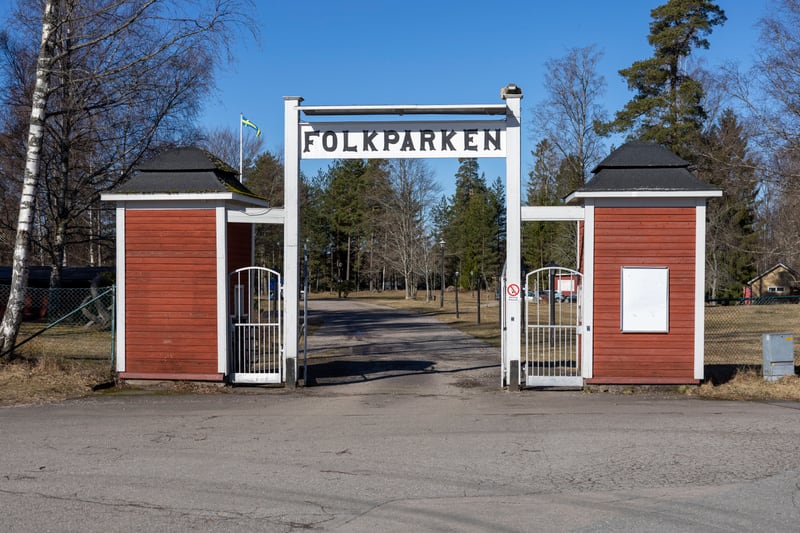 En bild tagen i dagsljus visar entrén till Folkparken. Ingången består av två röda byggnader med svarta tak och vita pelare som håller upp en skylt med texten ”FOLKPARKEN”. Mellan byggnaderna finns en asfalterad väg som leder in i parken. På varje si