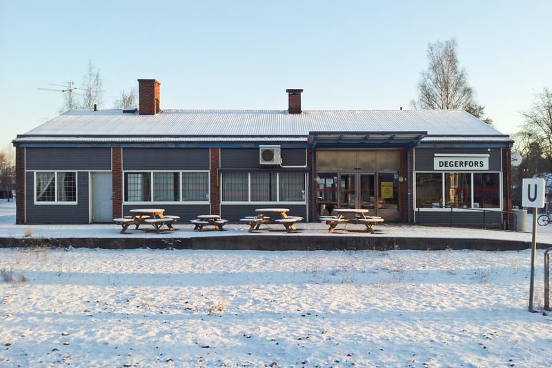 En enplansbyggnad med grå väggar och tegeldetaljer syns under ett snötäckt tak. På taket finns två skorstenar och en antenn. Utanför byggnaden, på en upphöjd plattform, står flera träbord med tillhörande bänkar, även de delvis täckta av snö. En del a