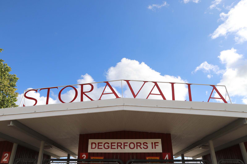 Bilden visar entréområdet till en arena eller idrottsplats, fotograferad nedifrån mot himlen. Ovanpå en röd byggnad med vitt tak och vita supports finns stora, röda bokstäver som stavar "STORAVALLA". Under taket, på den röda byggnadens framsida, sitt