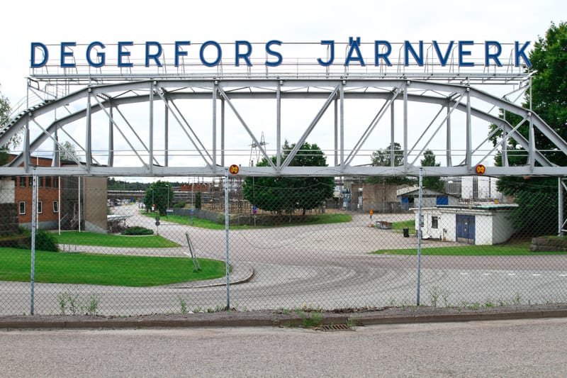 Ett högt, grått metallgaller med texten 'DEGERFORS JÄRNVERK' i blå versaler sträcker sig över en entré till ett industriområde. Under gallret syns en inhägnad med cyklonstängsel, och bakom den finns asfalterade vägar, gräsmattor och olika byggnader.
