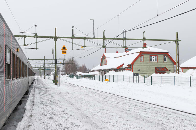 Bilden visar en järnvägsstation täckt av snö under en grå vinterdag. Ett tåg står vid en plattform till vänster, med delar av dess silverfärgade vagn synlig. Spåren är delvis snötäckta, och en plattform sträcker sig mot mitten av bilden. I bakgrunden