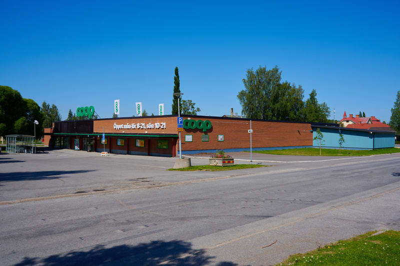 Bilden visar utsidan av en Coop-butik under dagtid med klarblå himmel. Butiken har en rödbrun tegelfasad och en mörkbrun del, med den gröna Coop-logotypen framträdande ovanför ingången. Mindre Coop-flaggor syns på taket. Vid ingången står en informat
