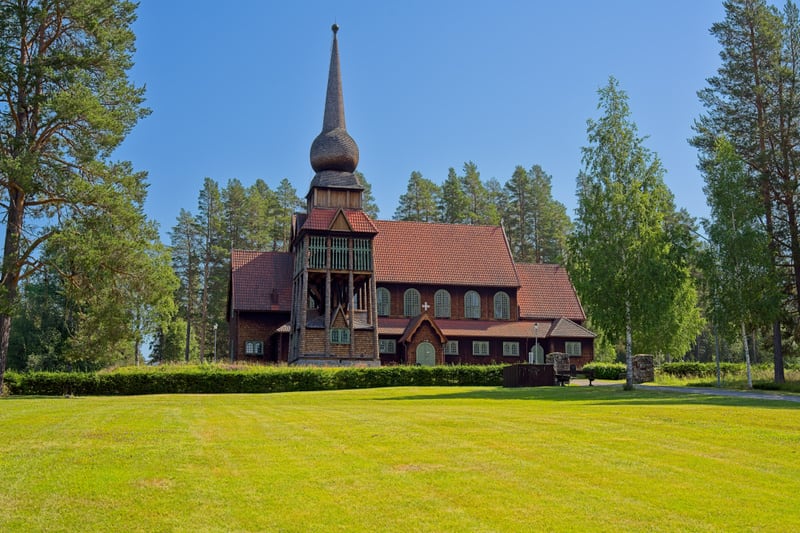Bilden visar en kyrkobyggnad i trä med rött tegeltak. Kyrkan har en distinkt klockstapel med en spira i förgrunden till vänster om mitt. Byggnaden har många fönster, några med rundade toppar. Intill kyrkan står flera grönskande träd, varav några är b